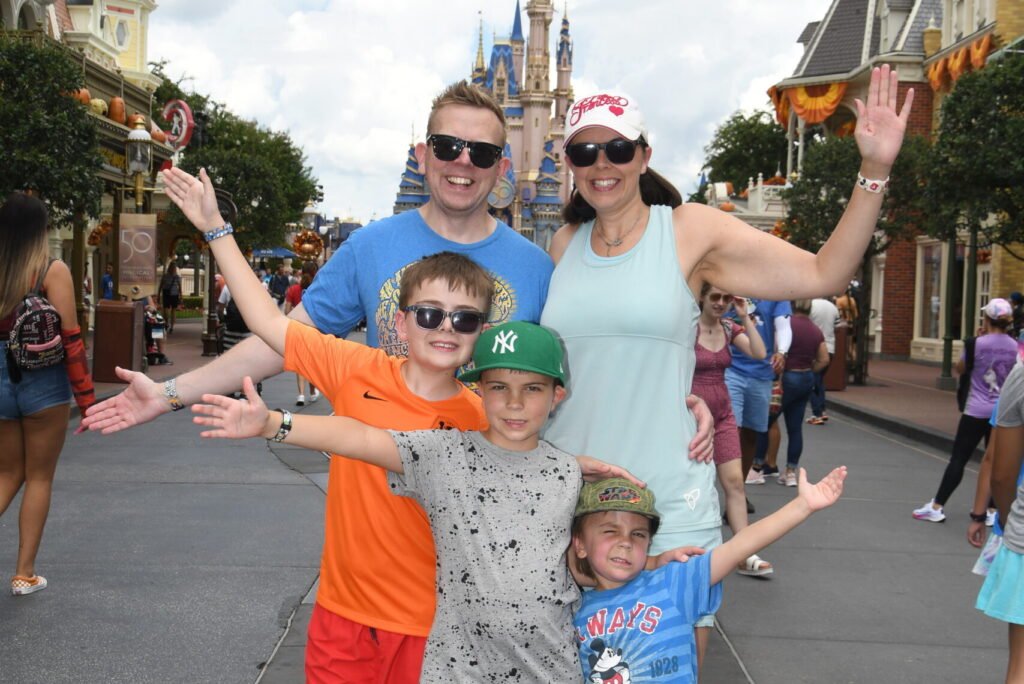 A smiling family stand in front of the magic family with their hands up. There are two adults and three boys.