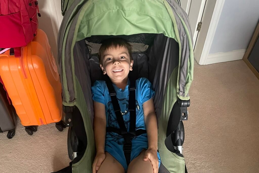 A boy of approximately 4 years old sits smiling in a pushchair in a house.