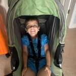 A boy of approximately 4 years old sits smiling in a pushchair in a house.