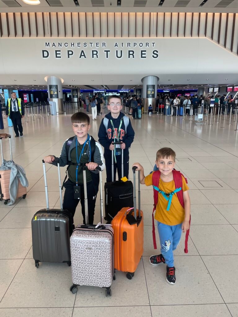 Three children stand holding cabin suitcase handles in their hands, smiling. They are waiting to go through to the departures area of the airport.