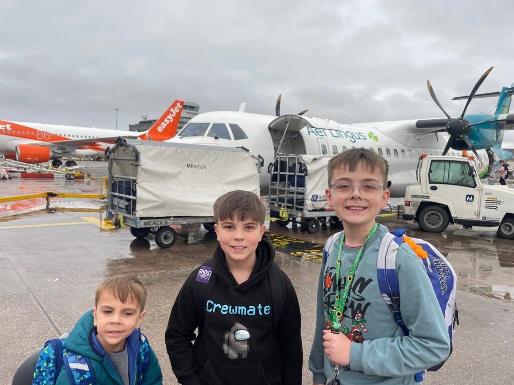 Three children stand smiling in front of a small Aer Lingus plane about to board.