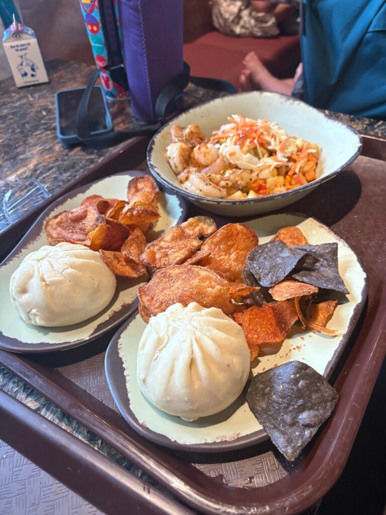 A tray shows three meals bought from a quick service in Disneys Animal Kingdom, two of which are bao buns with crisps, the other is a bowl of veggies
