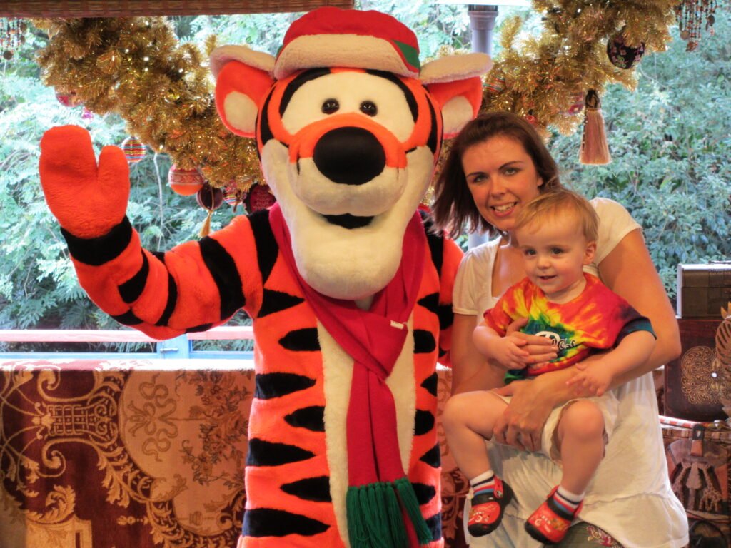 A mum holds a toddler next to Tigger dressed in Christmas hat at a Walt Disney World special event